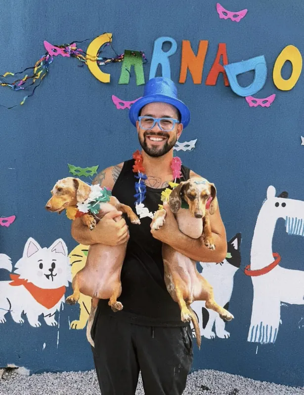 Vinícius fantasiado de Carnaval segurando dois Dachshunds com colares de flores